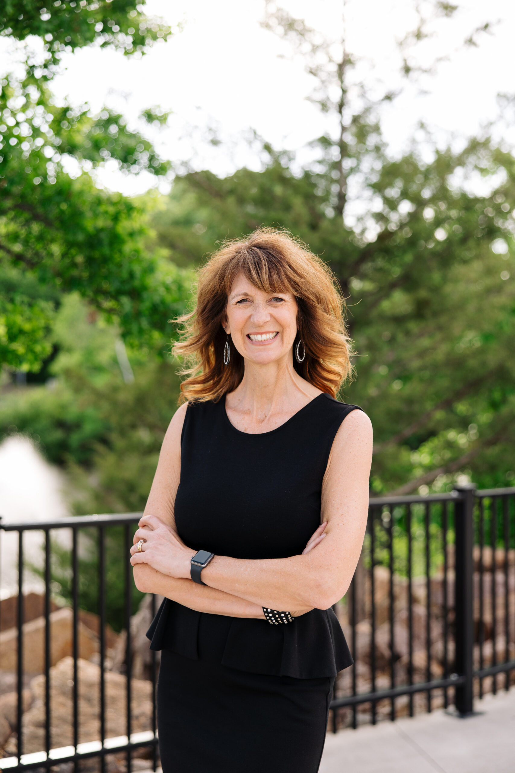 Smiling woman in a black sleeveless dress standing outdoors, arms crossed, with a black railing and greenery behind her.