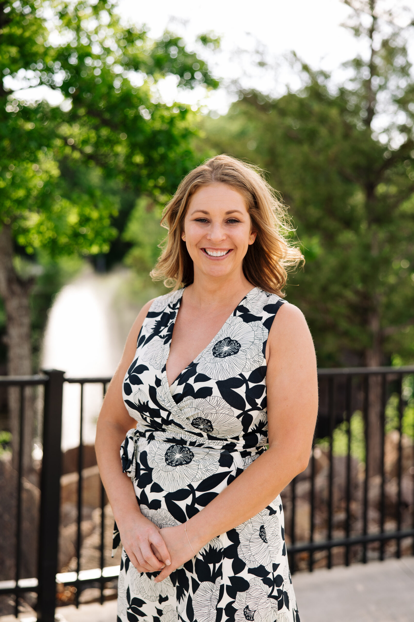 Smiling woman in a black-and-white floral dress standing outdoors near a black railing with green trees behind her