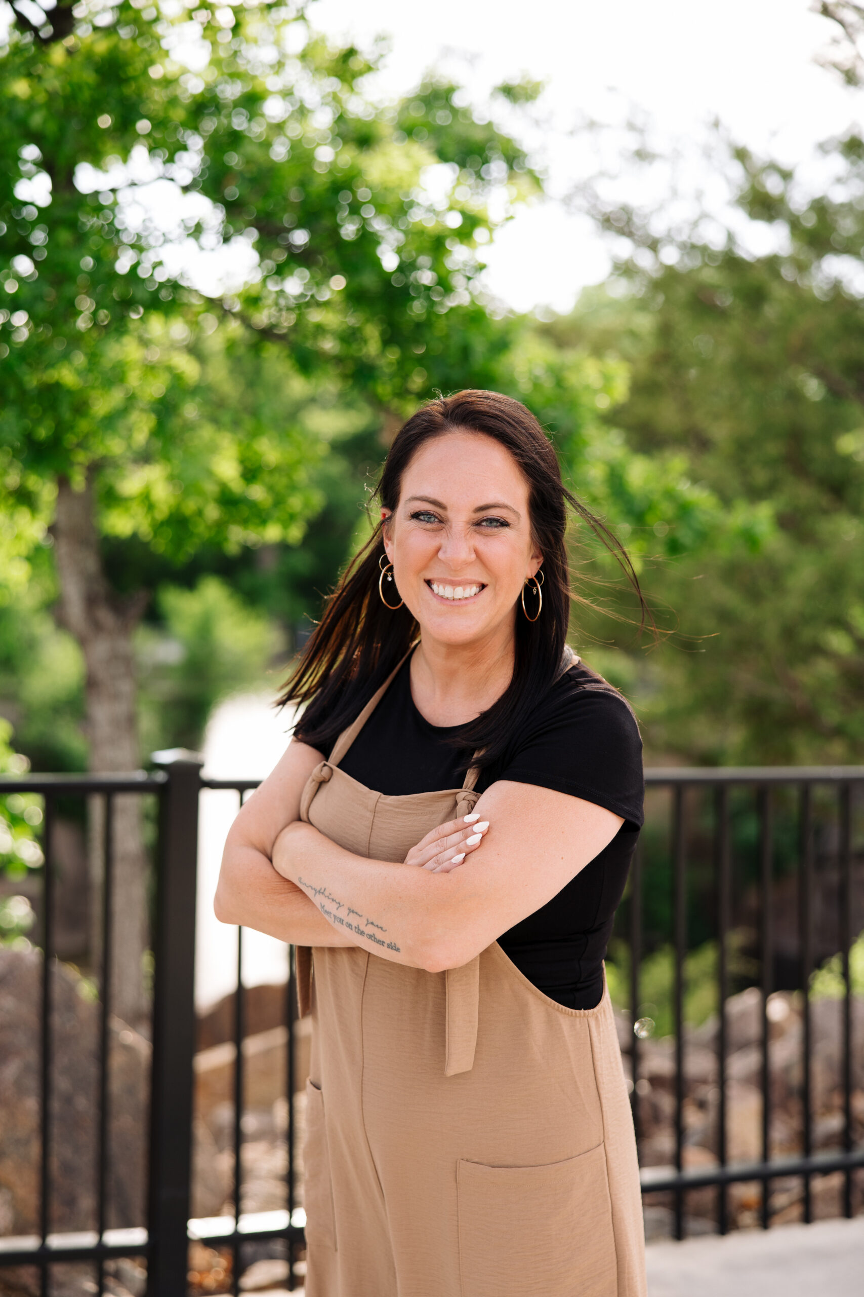 Smiling woman with brown hair and hoop earrings, arms crossed, wearing a black shirt and tan apron in a sunny park with a black railing behind her.