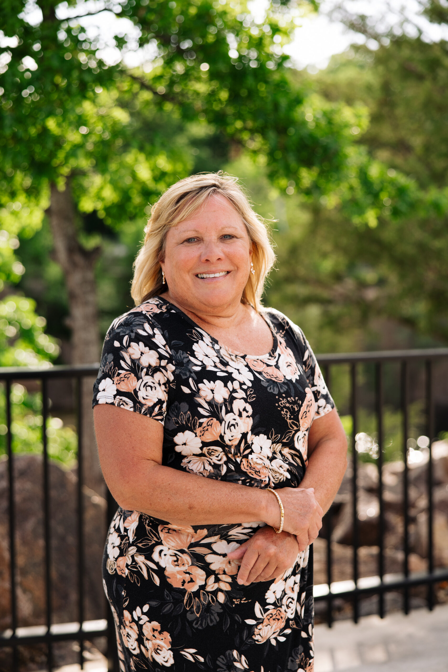 Smiling woman in a black floral dress standing outdoors with green trees in the background.