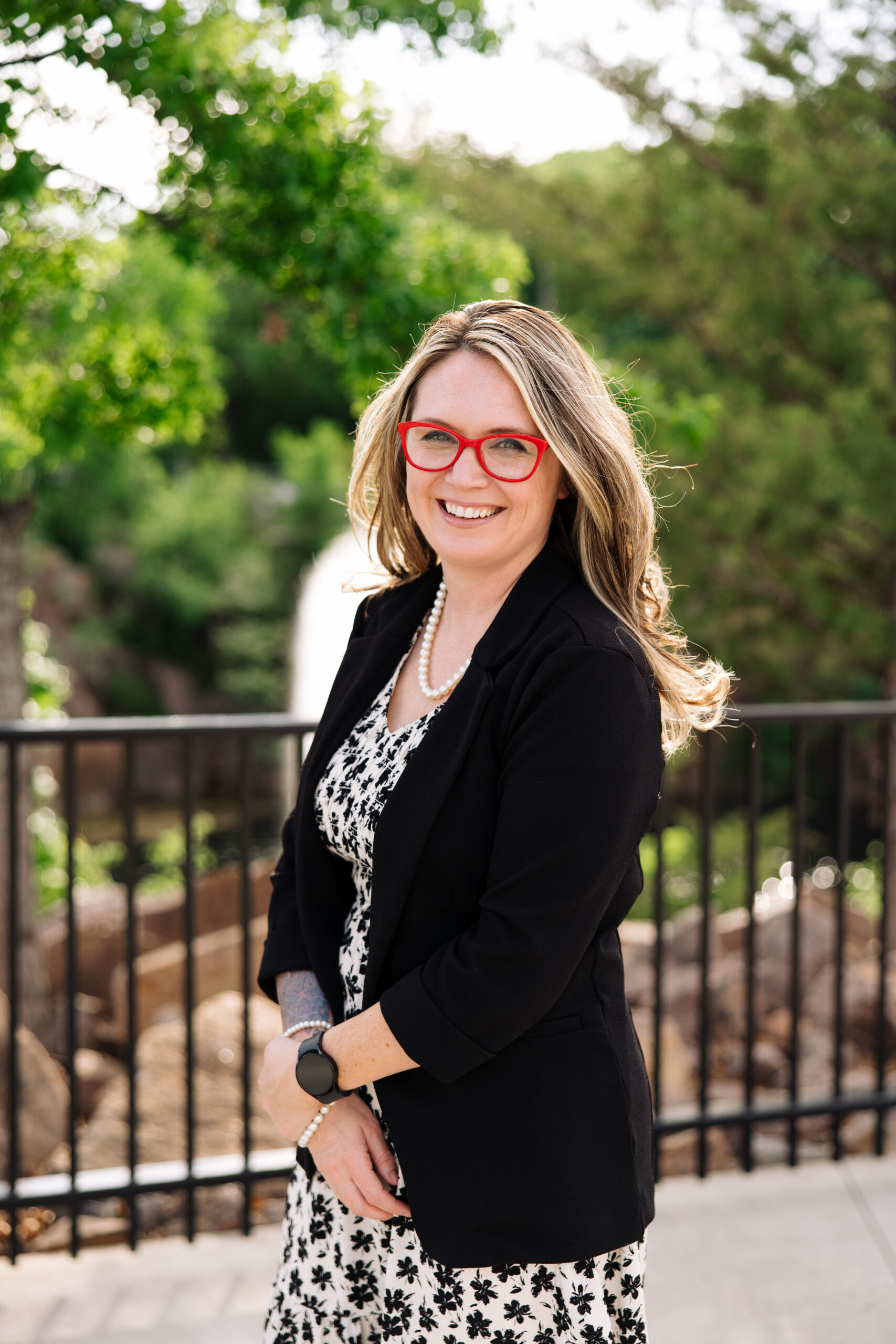 Smiling woman with red glasses, black blazer, pearl necklace, and floral dress outdoors by a railing.